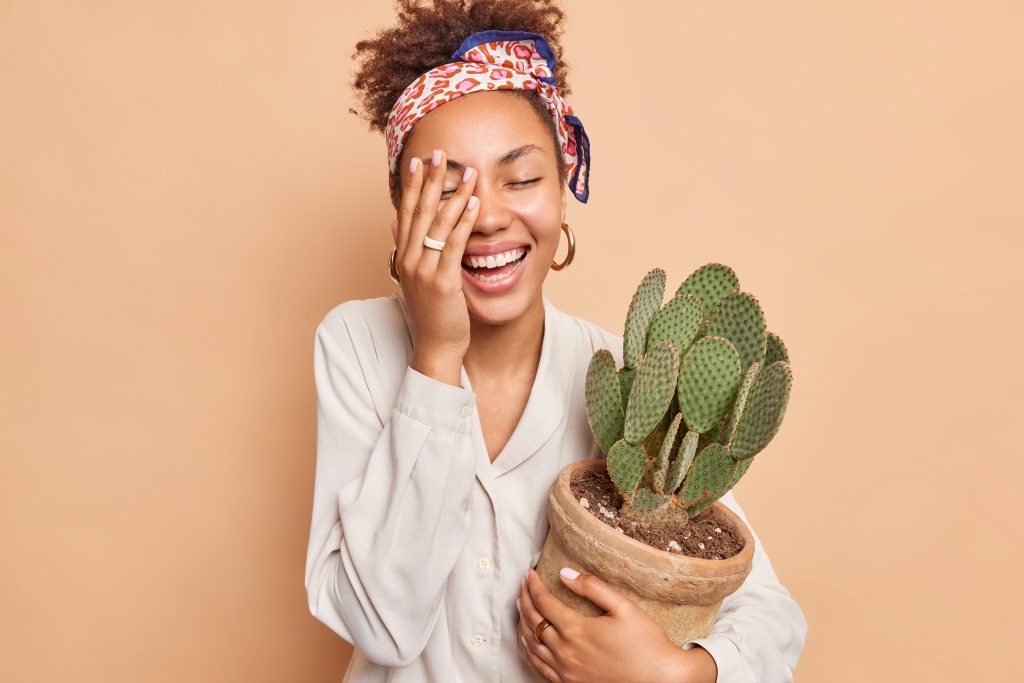 Cheerful dark skinned female model makes face palm smiles broadly has happy mood embraces pot with cactus wears white shirt and headkerchief isolated over beige background takes care of houseplants