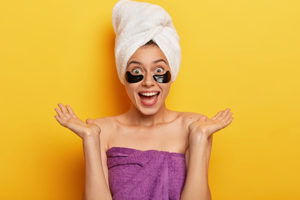 People, wellness and rejuvenation concept. positive european woman spreads palms sideways, makes beauty step for looking younger, wears cosmetic patches under eyes for reducing bags and puffiness