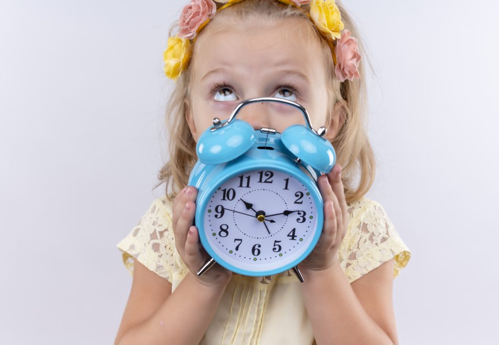 A lovely little girl wearing yellow shirt in floral headband showing blue alarm clock while looking up on a white background