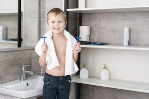 Portrait little boy with towel around his neck holding blue razor hands standing bathroom