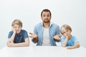 Portrait of questioned good looking european father sitting at table with bored and upset sons, shrugging with raised palms, being clueless how to raise boys alone, sitting confused over grey wall